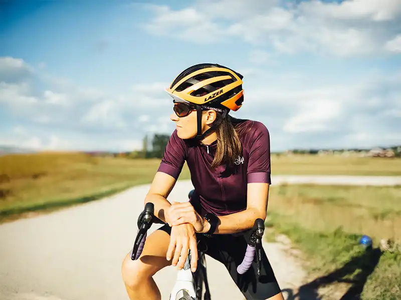 Radfahrerin mit dunkler Sportsonnenbrille und Helm, ruht sich auf einem Rennrad in ländlicher Umgebung aus.