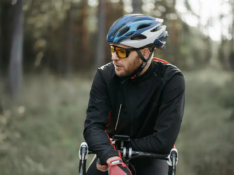 Männlicher Radfahrer mit Sportsonnenbrille mit gelben Gläsern und Helm, der sich in einem Waldgebiet auf dem Fahrrad ausruht.