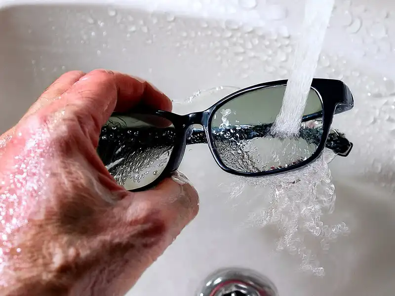 Eine Person spült Oakley-Sonnenbrillen unter fließendem Wasser in einem Waschbecken, mit Wassertropfen auf den Gläsern und dem Rahmen.