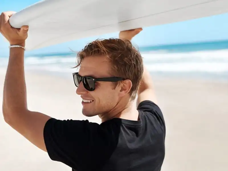 Mann mit Sonnenbrille und Surfbrett an einem sonnigen Strand