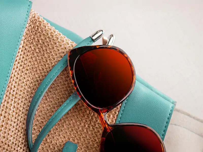 Sonnenbrille mit braunem Rahmen und burgunderfarbenen Gläsern auf einer Handtasche aus Stroh und Blaugrün.