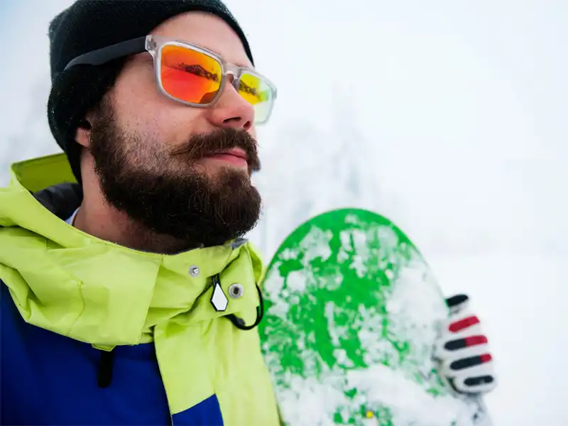 Hombre con ropa de invierno usando gafas deportivas espejadas, sosteniendo una tabla de snowboard verde en un paisaje nevado.