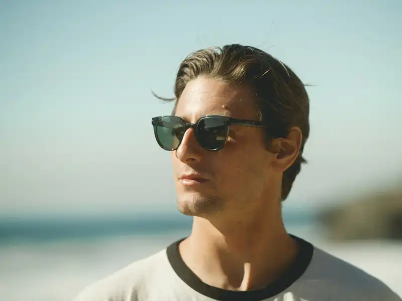 Hombre mirando de lado con gafas de sol, mostrando cómo protegen los ojos del resplandor solar cerca del mar.