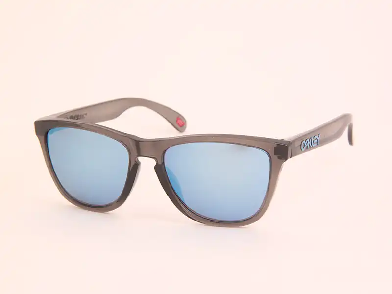 Unas modernas gafas Oakley tipo shield sobre una valla de madera, evocando el diseño de las originales Factory Pilot Eyeshades de 1984, las primeras gafas de rendimiento de la marca.