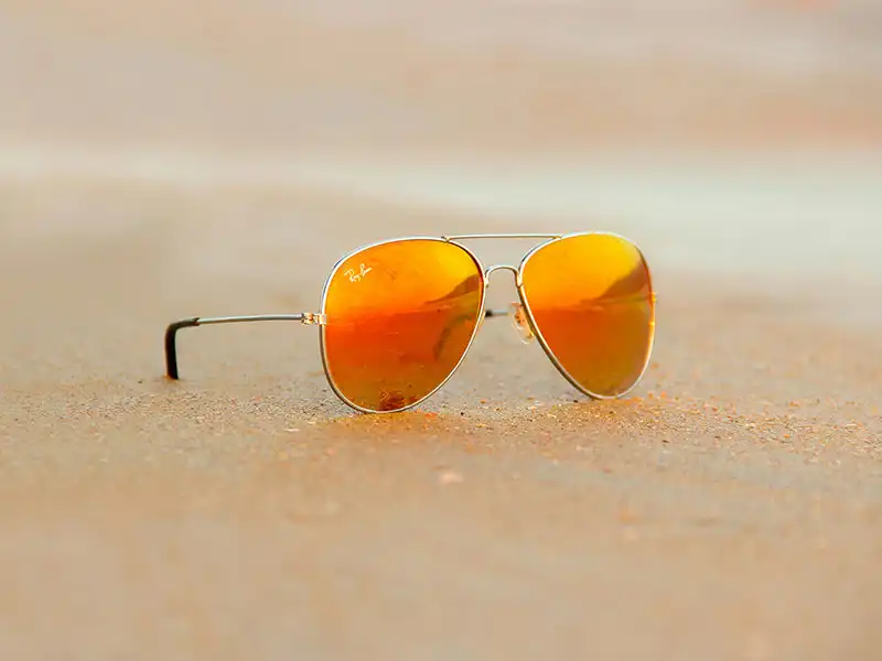 Lunettes aviator métalliques dorées avec verres miroirs orange posées sur du sable, représentant un style classique et emblématique Lunettes aviator métalliques dorées avec verres miroirs orange posées sur du sable, représentant un style classique et emblématique