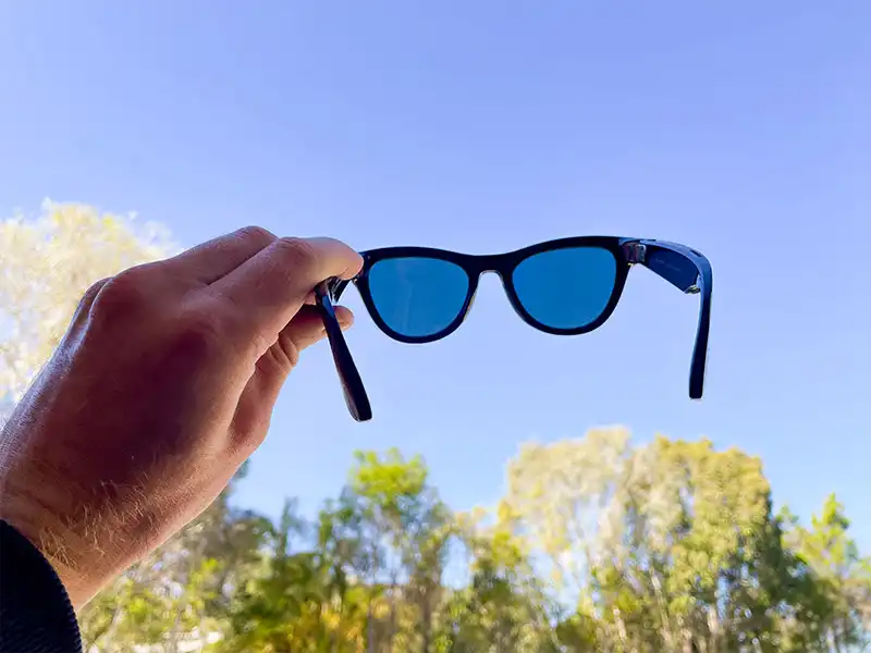 Hand hält eine Ray-Ban Meta-Sonnenbrille mit blauen Gläsern vor einem klaren Himmel und Bäumen