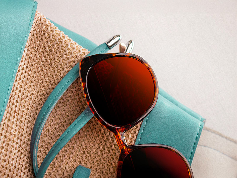 Brown-framed sunglasses with burgundy lenses placed on a straw and teal handbag.