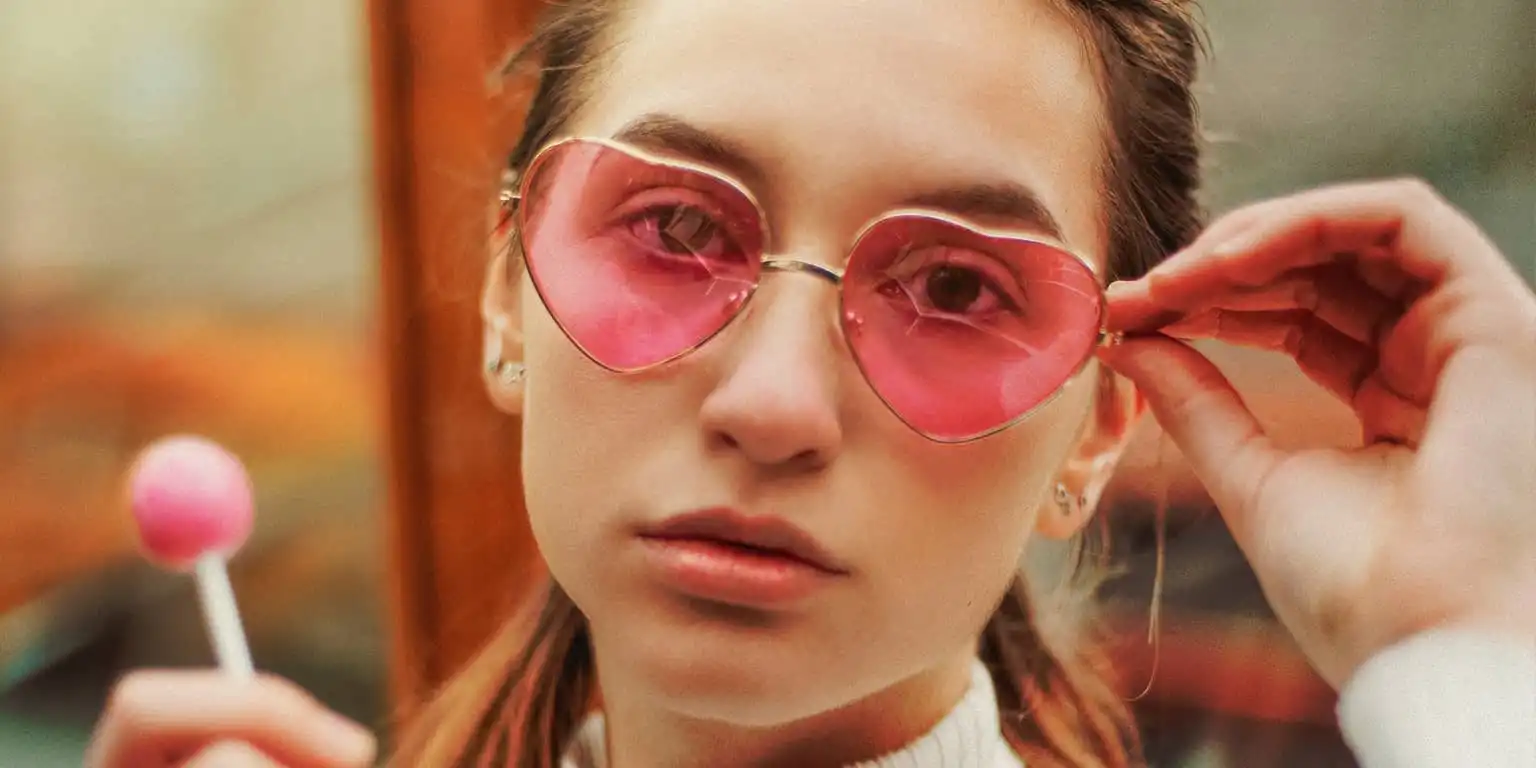 A girl in rose tinted loveheart shaped sunglasses with a lollipop