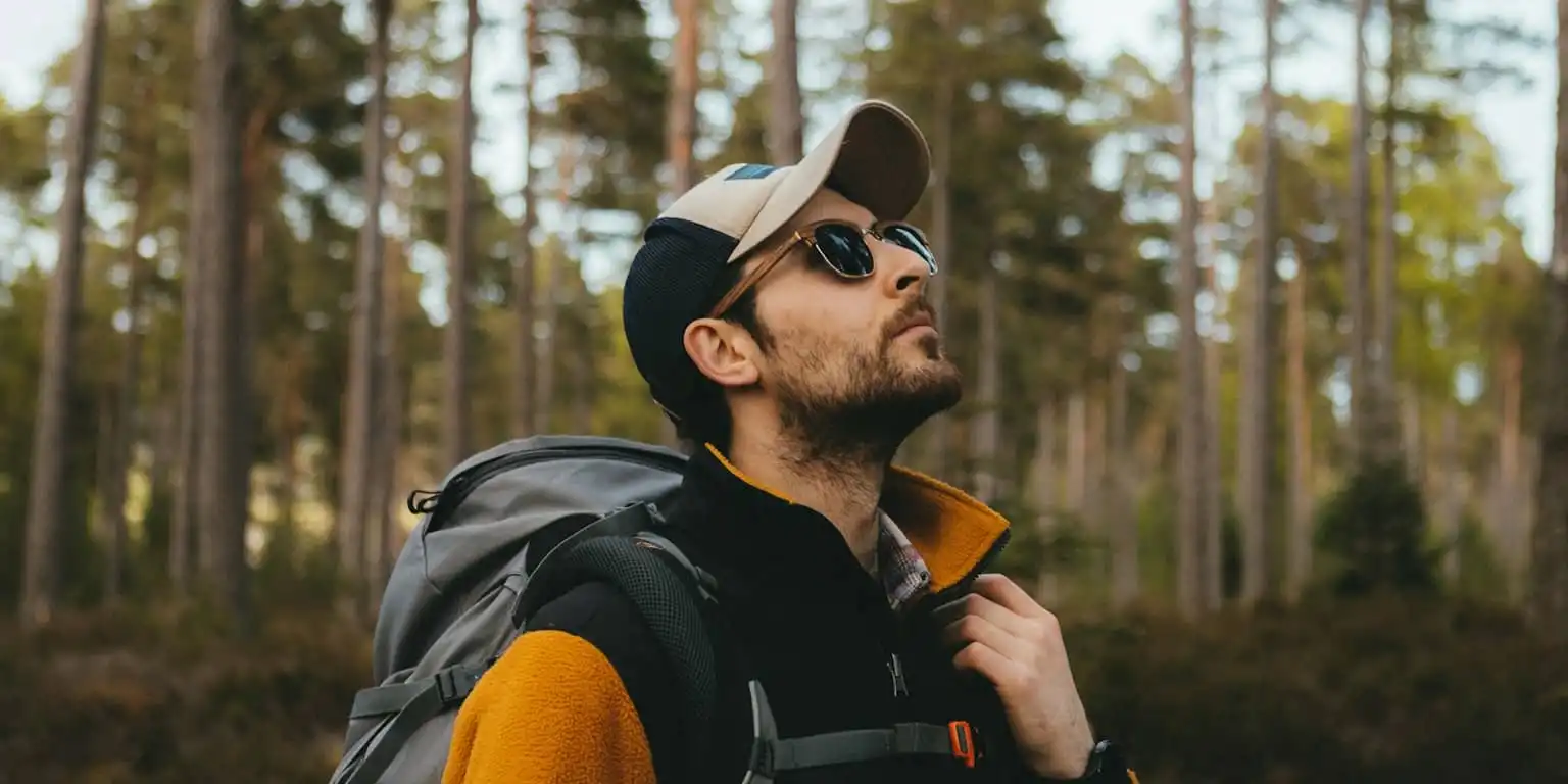 A man looking up in the forest wearing a cap and brown framed sunglasses